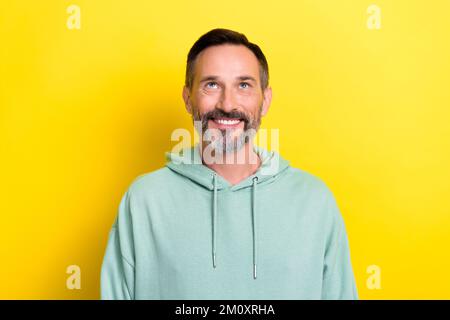 Portrait of mature age brunet hair retired man wear pastel green hoodie smiling look up empty space good news isolated on yellow color background Stock Photo