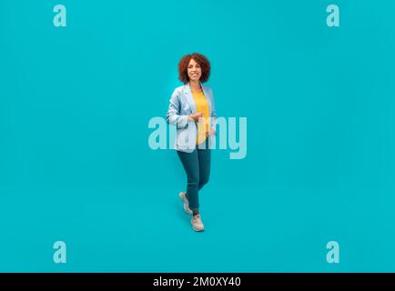 Smiling successful African American woman with curly hair in jacket on blue background in full growth Stock Photo
