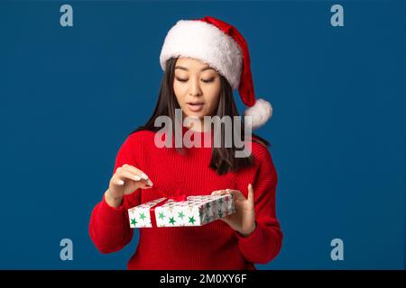 Pleasantly excited woman unpacking christmas present. Beautiful woman in santa claus hat on blue background Xmas gift Stock Photo
