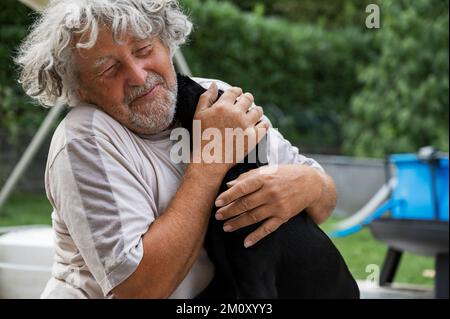 Young black labrador puppy kissing a cheerful senior man with grey curly hair outside in backyard. Stock Photo