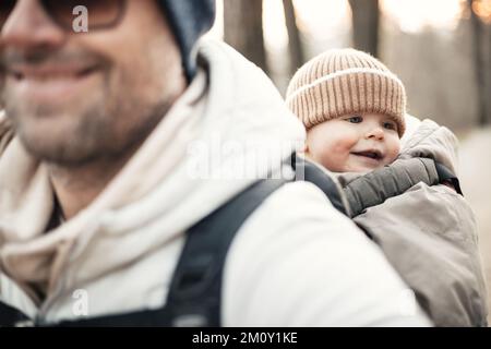 Sporty father carrying his infant son wearing winter jumpsuit and cap in backpack carrier hiking in autumn forest Stock Photo