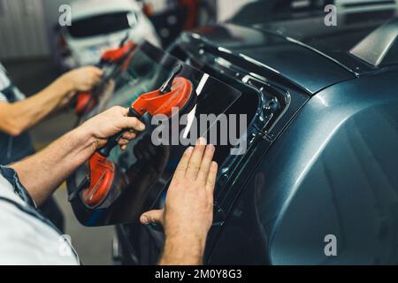 Automobile repair process. Closeup indoor portrait of two unrecognizable caucasian blue collar workers using suction cups to remove rear window from a black car. High quality photo Stock Photo
