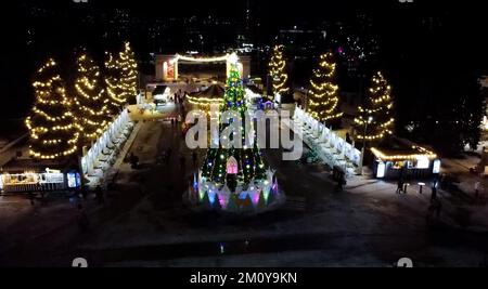 Christmas tree decorated decorative luminous garlands and children's spinning carousel in park in city on winter night. People walking in park. New Year Christmas Stock Photo