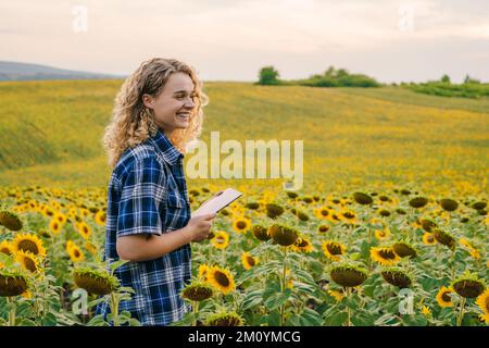 Woman farmer using tablet for statistics collection, standing in the sunflower field. Analyzing agricultural data with intelligent technology. Smart Stock Photo