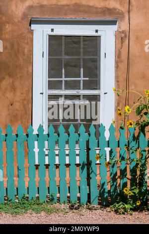 Vertical view of turquoise color wood fence and sunflowers set in front of a window and rustic adobe wall in Santa Fe, New Mexico Stock Photo