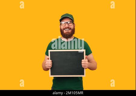 Happy bearded man wearing courier uniform and glasses is holding a blackboard over yellow background. Stock Photo