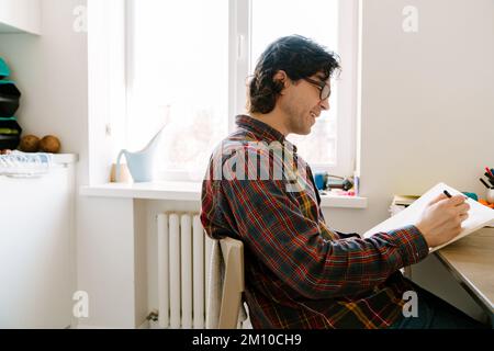 White man laughing and writing down notes while sitting in kitchen at home Stock Photo