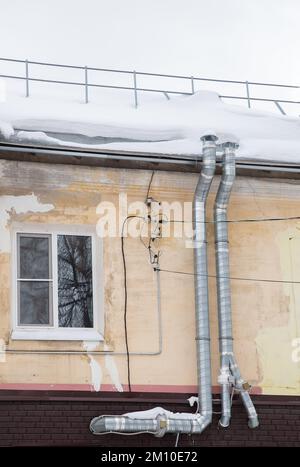 Two long metal exhaust pipes for ventilation on the wall of an old house. The roof of the building in white snow. Cloudy, cold winter day, soft light. Stock Photo