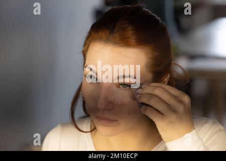 Tired and stressed girl in front of the mirror doing her make up before going to work. A sad look out of boredom from the routine and the mask worn to Stock Photo