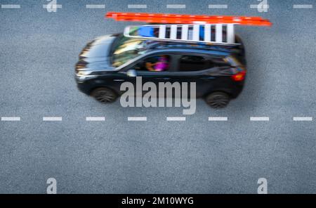 Top view of a driver going to work in a compact black car with an orange step ladder strapped to the car roof rack. Stock Photo