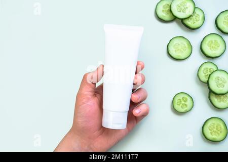 Mockup of white squeeze bottle plastic cosmetic tube for branding in Caucasian man's hand and fresh cucumber slices on green background. Unbranded lot Stock Photo