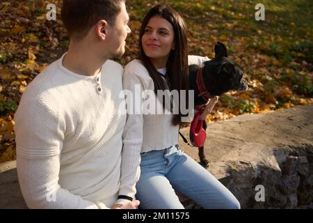 Joyful woman, guy and black dog are sitting hugging on parapet Stock Photo