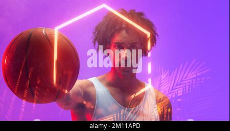 Portrait of biracial basketball player with afro hair holding ball by illuminated hexagon and plants. Purple, serious, copy space, composite, sport, c Stock Photo