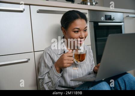 Cheerful African woman is using laptop in the kitchen Stock Photo