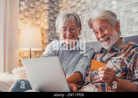 Portrait of couple of cute two happy and excited seniors buying and spending money shopping online with laptop on the sofa at home. Using credit card together buying on internet. Stock Photo