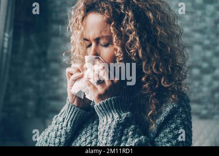 Side view of woman blowing nose for flu influenza symptoms in winter at home. Temperature reduction indoor to save energy gas costs. Female people with virus contagion using paper tissues. Disease Stock Photo