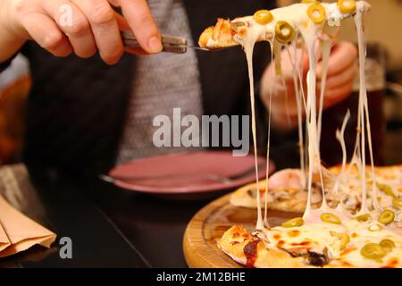 Defocus female hand holding slice of italian pizza Tortilla with grated Mozzarella. Pizza with olives, Basil and cheese close up. Pizzeria outside Stock Photo