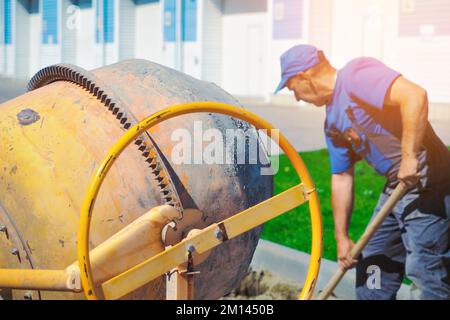 Uniformed construction worker works on construction site on summer day. Elderly bricklayer shovels cement and sand into concrete mixer. Authentic workflow.. Stock Photo
