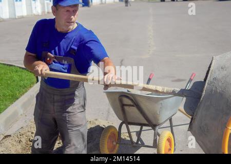Builder with shovel interferes with cement mortar for construction. Elderly working man works on construction site on summer day.. Stock Photo