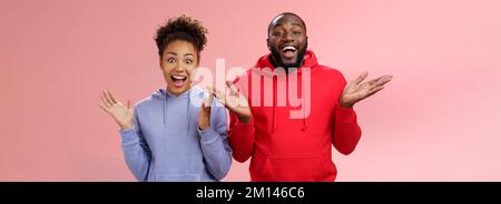 Guys receive nice surprise. Two attractive carefree surprised happy african man woman clapping hands joyfully greeting welcoming guests inviting Stock Photo