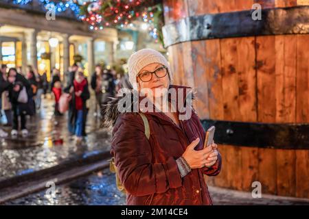 Happy senior woman tourist in a knitted hat, jacket, with a backpack walks along the street in a rainy festive city, smiling, and used phone. Christmas lights lights and crowd in the background. Stock Photo