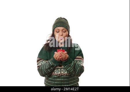 Stunning woman in wool hat and green knitted sweater, makes wish and blows out flame on red candle, on white background Stock Photo
