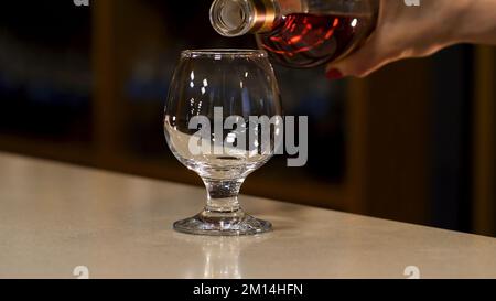 Close up of woman hand pouring cognac to the glass standing on the bar counter. Hand pouring alcohol into a glass on the blurred background of a dark Stock Photo