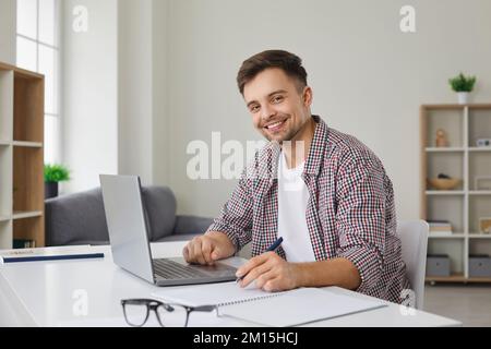 Portrait of happy man sitting at desk, working on laptop computer and taking notes Stock Photo