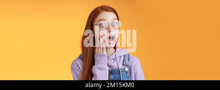 Shocked impressed speechless young redhead sensitive european girl glasses wearing hoodie dungarees drop jaw gasping astonished look left wide eyes Stock Photo
