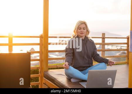 A calm woman sits on a sun lounger near the sea, gets distracted from working at the computer, meditates and removes negative emotions, combines Stock Photo