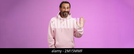 Cheeky charismatic funny happy smiling mature man bearded grey hair in pink hoodie waving palm hello nice meet you greetng gesture welcoming guests Stock Photo