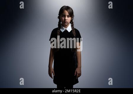 Portrait of little girl with Wednesday Addams costume during Halloween. Serious expression and dark atmosphere with dark background. Stock Photo