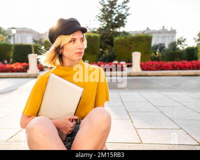 Pensive young female freelancer with blond hair in casual clothes and hat sitting on stairs in city park with laptop in hand and looking away Stock Photo