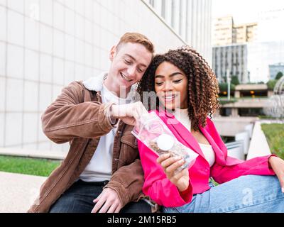 Happy multiracial students in street style clothes sitting on bench close to each other near university building and eating nuts Stock Photo