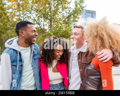 Group of cheerful multiracial friends in street style outfits walking in park near modern buildings having fun together Stock Photo