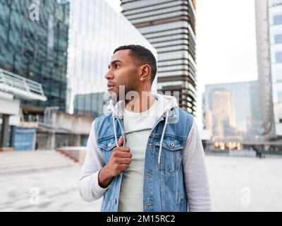 Confident young Hispanic male millennial in casual hooded jacket standing on street against modern buildings and looking away Stock Photo
