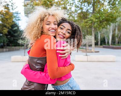 Smiling young female students in colorful stylish outfits with curly hair standing in urban city park and embracing while looking at each other Stock Photo