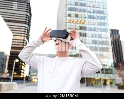 Amazed young male millennial in casual clothes watching video in VR goggles with opened mouth while standing on city street against modern skyscrapers Stock Photo