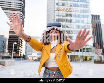 Delighted young African American woman in stylish yellow jacket smiling while standing on city street experiencing virtual reality in modern headset Stock Photo