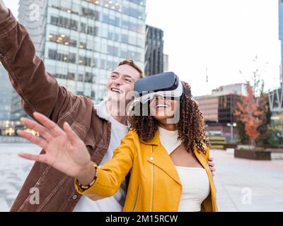 Delighted young man in stylish jacket smiling and pointing away while standing on city street near ethnic female friend experiencing virtual reality i Stock Photo