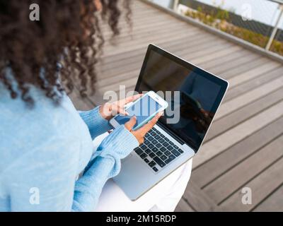 From above back view of cropped unrecognizable Hispanic female student in blue turtleneck with Afro hairstyle sitting on wooden bench and chatting via Stock Photo