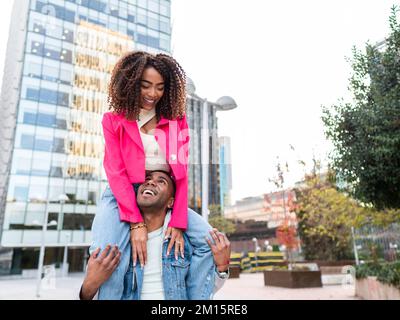 Cheerful young ethnic male millennial in casual clothes smiling happily while giving shoulder ride to delighted girlfriend against modern buildings on Stock Photo