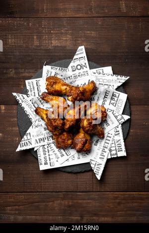 Top view of appetizing roasted chicken wings placed on black plate decorated with newspaper scraps on wooden table Stock Photo