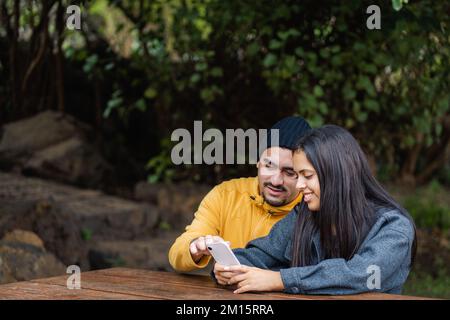 Hispanic man in outerwear pointing at smartphone screen for glad girlfriend while sitting at table on weekend day at park Stock Photo