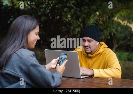Happy Hispanic woman browsing smartphone near man making video call via laptop while sitting at wooden table in park together Stock Photo
