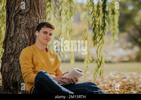 Smiling male in casual clothes in earphones sitting looking at camera on fallen leaves under willow tree and reading book in park Stock Photo