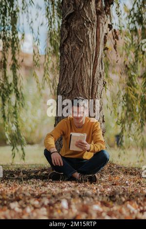 Full body of young male in casual clothes in earphones sitting on fallen leaves under willow tree and reading book in park Stock Photo
