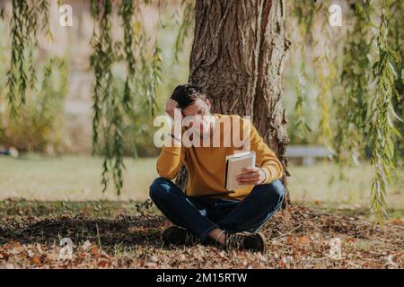 Full body of young thoughtful male in casual clothes in earphones sitting on fallen leaves under willow tree and reading book in park Stock Photo