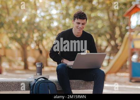 Concentrated smiling male student in casual outfit sitting on stone bench near thermomug while working remotely via laptop in autumn park Stock Photo