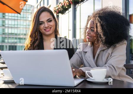 Elegant diverse women in coats having coffee while meeting in cafe in city downtown and browsing laptop discussing business Stock Photo